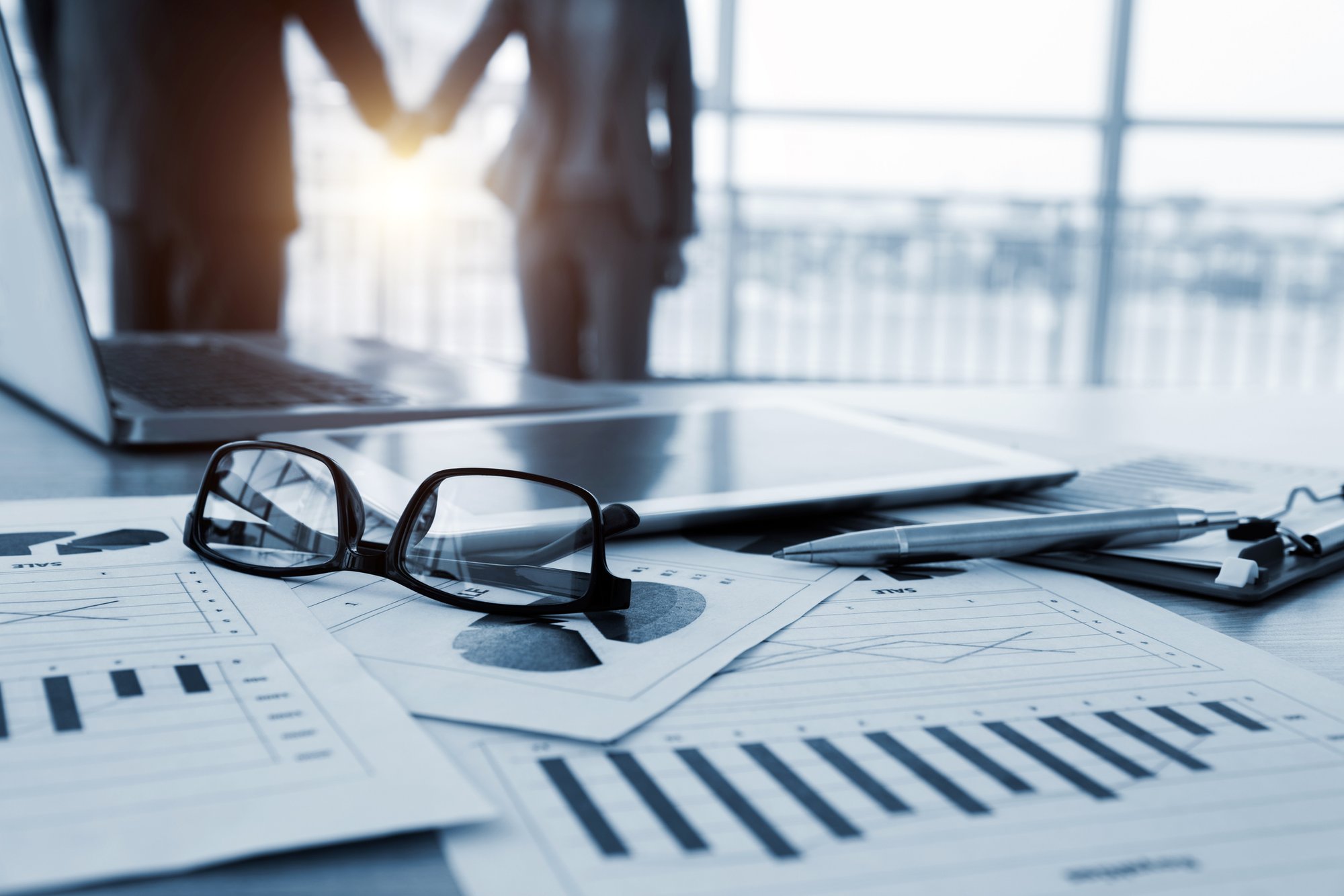 Business documents, glasses, and a laptop on a desk with two executives shaking hands in the background