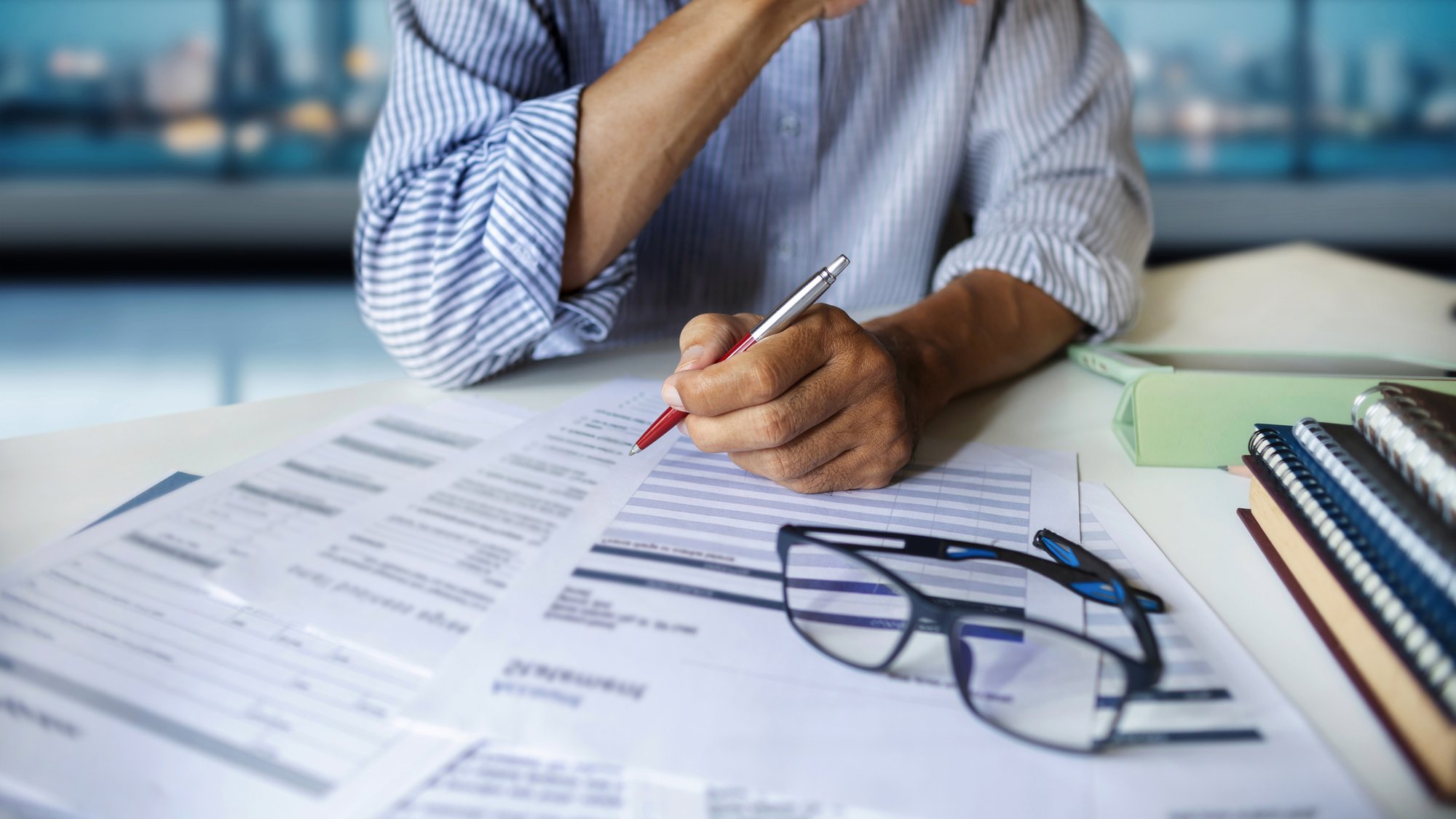 Close-up of a hand reviewing spend reports with a pen, glasses resting on the documents